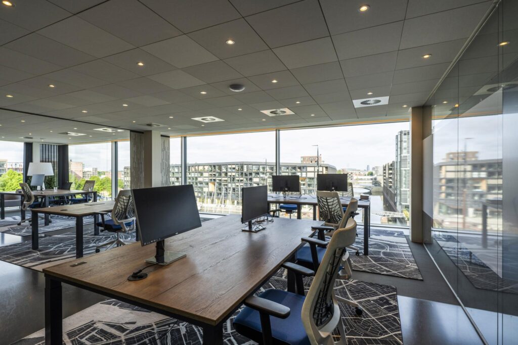 Modern open office space with large desks, ergonomic chairs, and a panoramic view of Krijn Taconiskade through floor-to-ceiling windows.